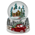 A festive Christmas snow globe figurine featuring a small snow-covered cabin, evergreen trees, a snowman, a red pickup truck carrying Santa, and a snowy base on a white background.