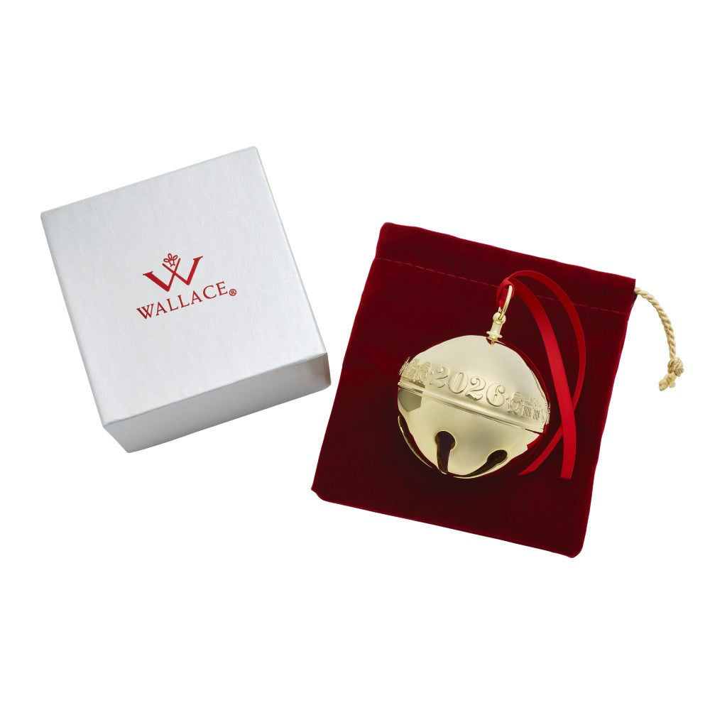 A gold-toned 2026 Christmas ornament shaped like a jingle bell, displayed next to its red velvet storage pouch and a white Wallace gift box on a white background.
